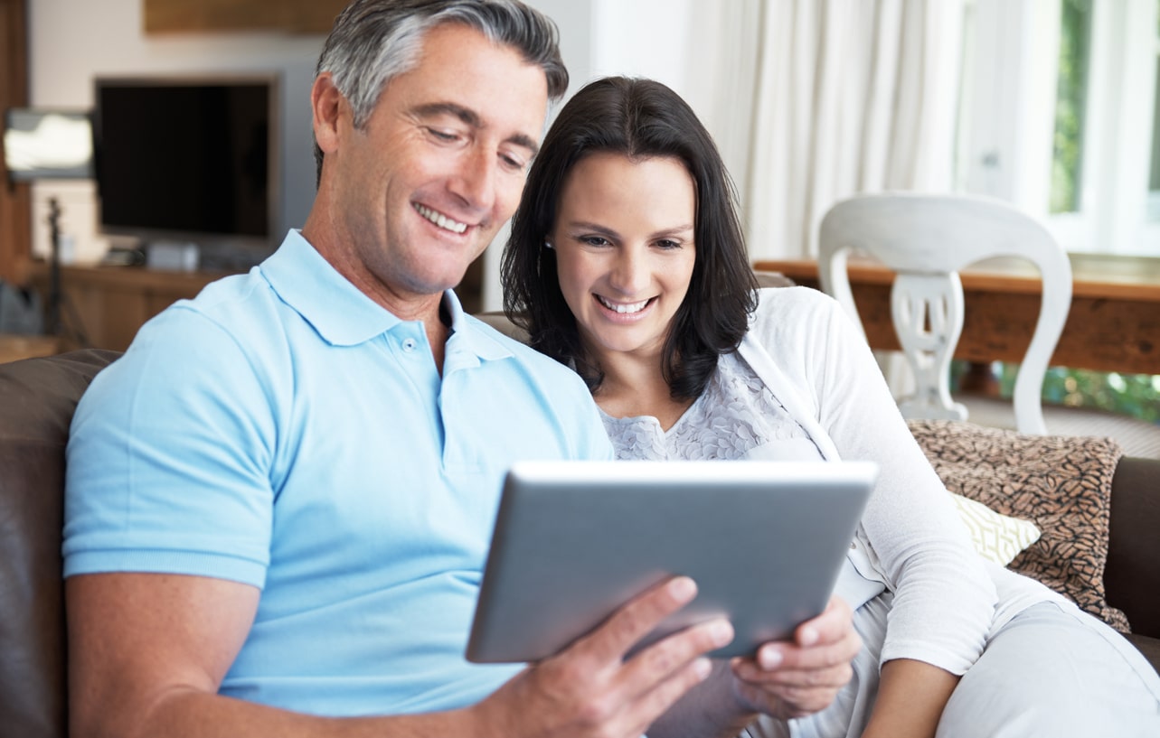 Middle-aged man and woman sitting on a couch smiling while using a tablet together