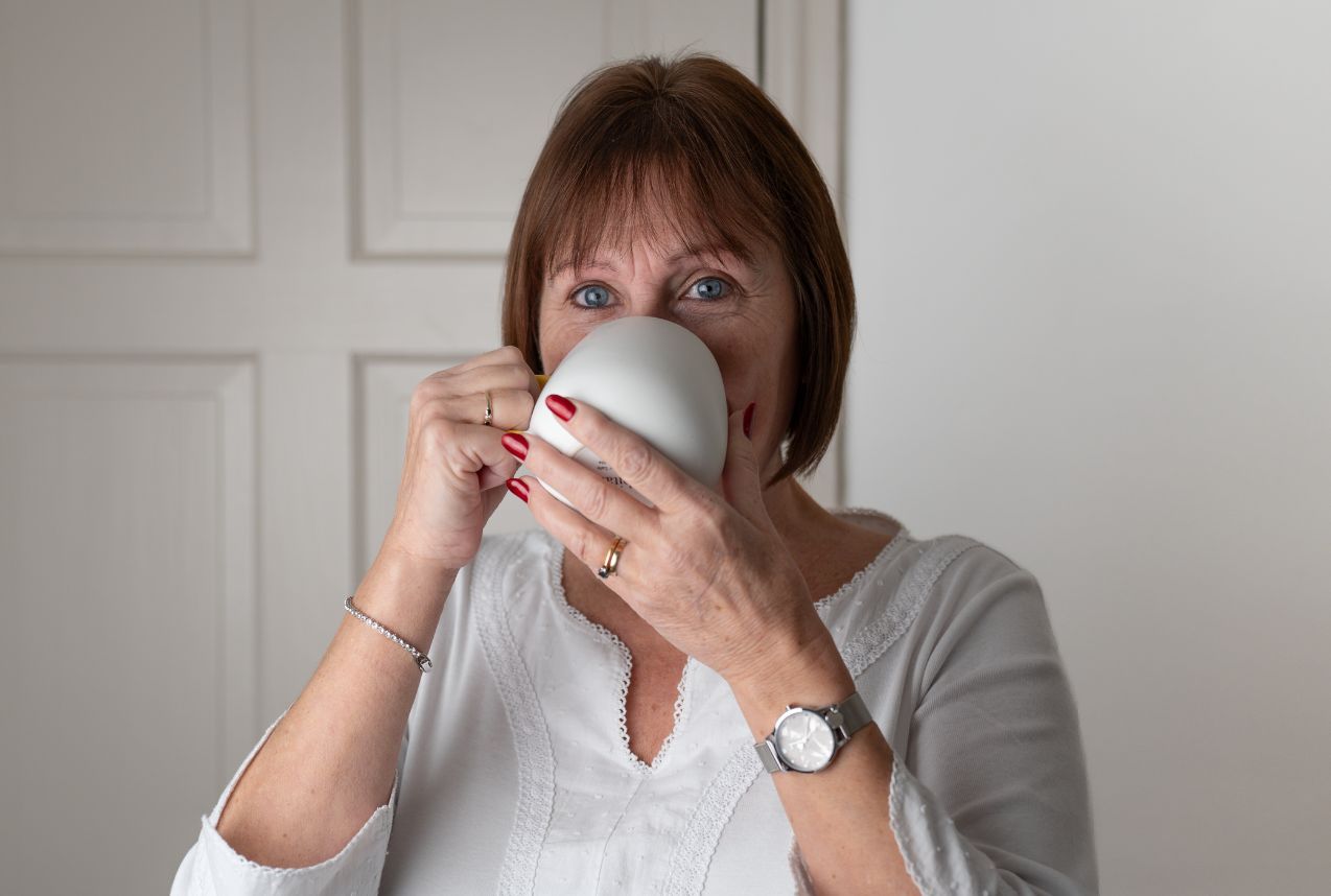 Woman with red nail polish sipping from a white mug, wearing a white blouse, silver watch and rings.